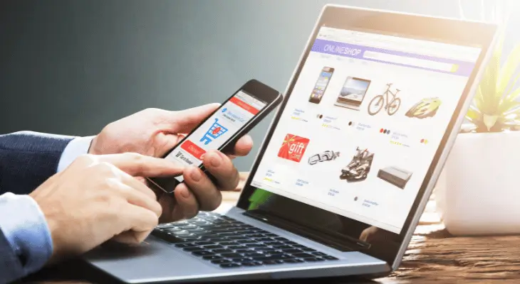 Imagem de homem mexendo em um app de e-commerce e um computador aberto em e na tela, uma página de loja virtual