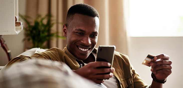 Homem olhando o celular com semblante de satisfação e segurando um cartão se crédito