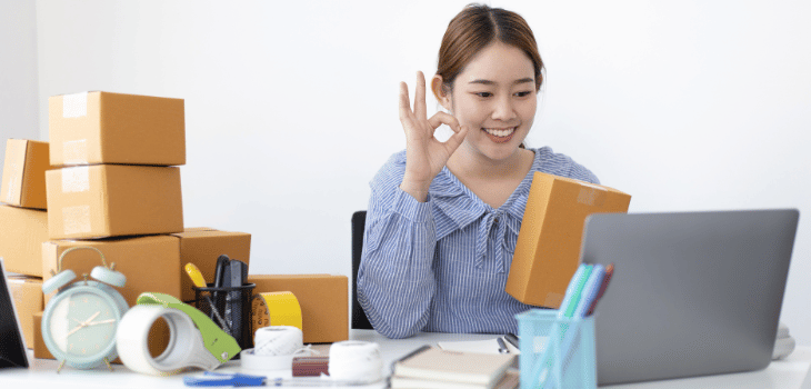 Imagem mostra mulher branca feliz na frente do notebook, com uma caixa de encomenda  em uma das mãos e atrás também há caixas.