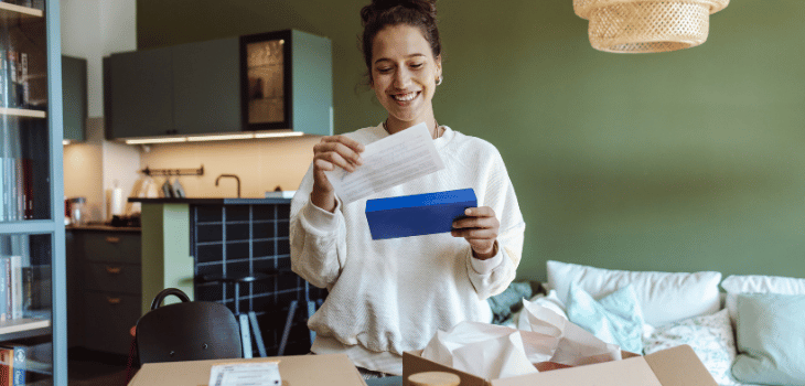 Mulher branca em pé, com caixas de encomendas em cima da mesa e lendo com o semblante feliz uma carta de de um dos pacotes