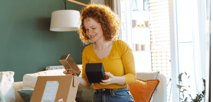 Mulher branca, ruiva, de camiseta amarela, sorrindo para os produtos recém recebidos