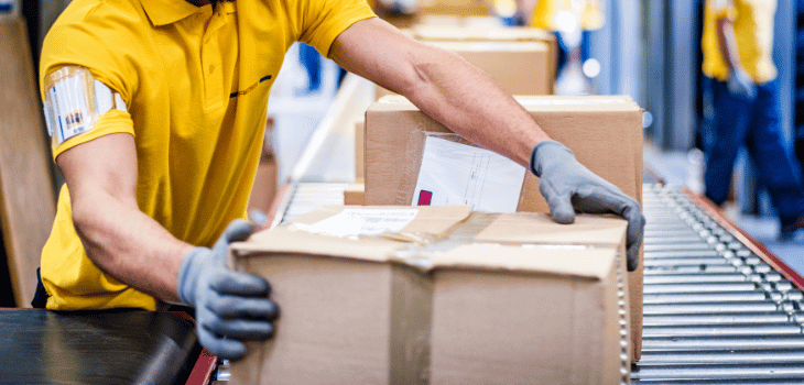 Homem usando uma camiseta amarela de uniforme, de luvas, segurando uma caixa de encomenda feita de papelão, em uma esteira de embalagens. 