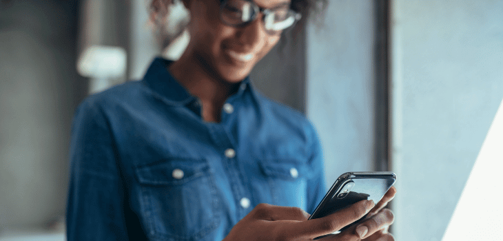 Mulher negra, vestindo uma camisa azul escuro, sorrindo enquanto olha a tela do celular.