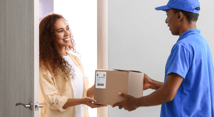 Mulher recebendo uma  caixa de papelão de encomenda de um entregador. Ambos são negros. Ela veste uma camisa estilo social rosa e blusa básica branca por baixo. Ele, o uniforme de uma empresa de cor azul, contendo boné da mesma cor. 