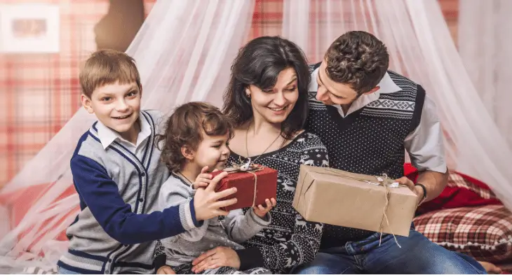 Imagem mulher recebendo presentes dos filhos e de um home que parece ser seu marido. Ela está sentada no meis, entre as crianças e o homem.