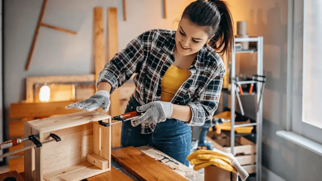 Imagem de mulher construindo uma caixa de madeira. Ela é  branca e está usando luvas e uma cerra.